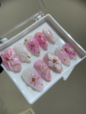 A set of ten pink and white false nails with floral and other designs. The nails are arranged in a white box.