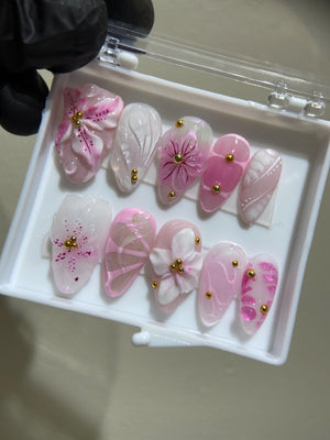 A set of ten pink and white false nails with floral designs and gold accents. The nails are arranged in a clear plastic case.