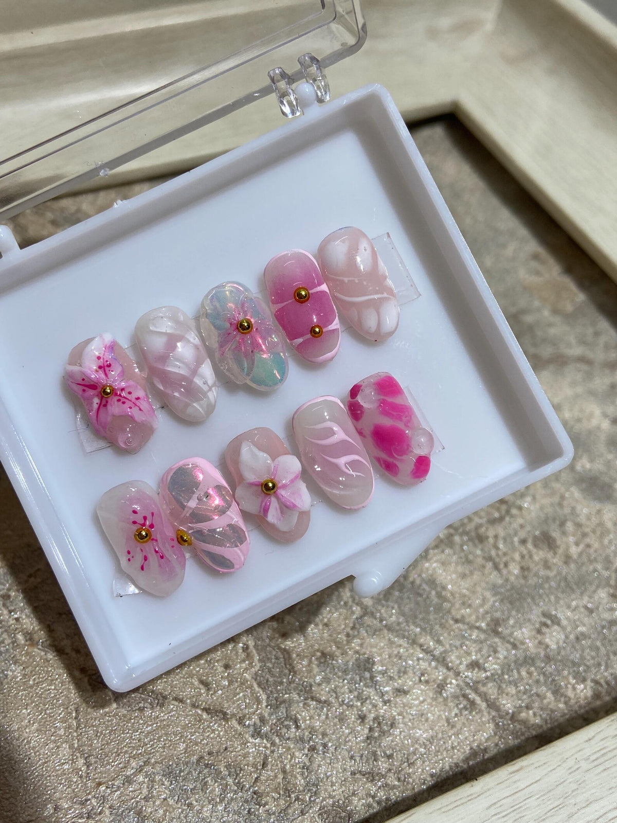 A set of eight pink and white press-on nails with floral designs and gold accents. The nails are arranged in a white box.
