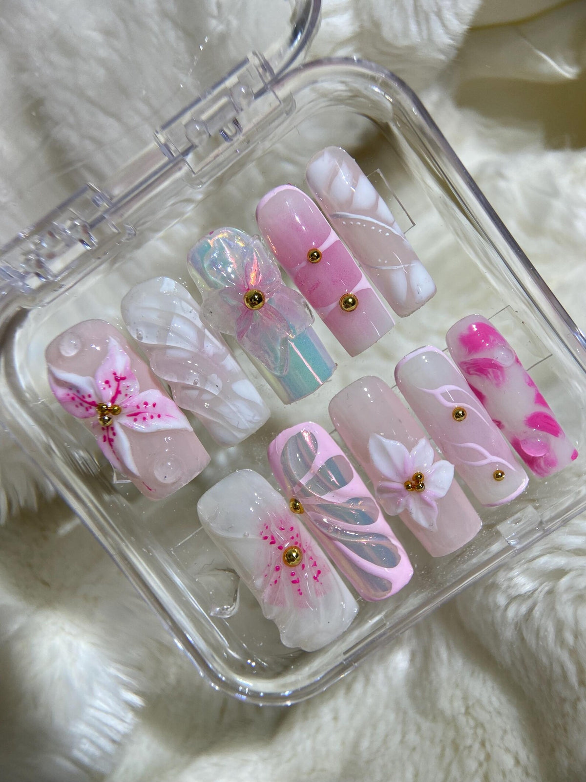 A set of ten pink and white artificial nails with floral designs and gold accents. The nails are arranged in a clear plastic case.