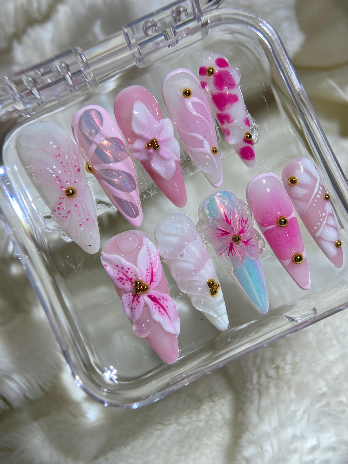 A set of ten pink and white artificial nails with 3D floral designs and gold accents. The nails are arranged in a clear plastic box.