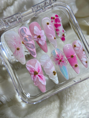 A set of ten pink and white artificial nails with 3D floral designs and gold accents. The nails are arranged in a clear plastic box.