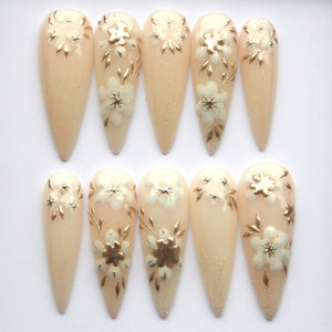 A set of ten artificial nails with a light beige base and gold glitter. Each nail features a white flower design with gold accents, including petals and leaves. The nails are almond-shaped.