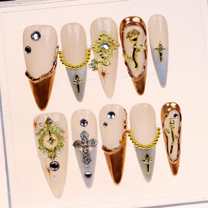 Set of ten false nails with a nude base and gold and silver embellishments. The embellishments include crosses, flowers, chains, and rhinestones.