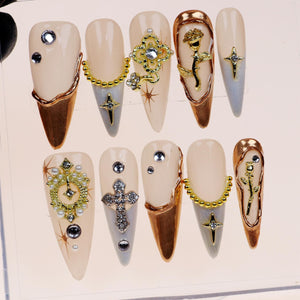 Set of ten false nails with various gold and silver embellishments, including crosses, flowers, and chains. The nails are a light pink color and have a long, pointed shape.