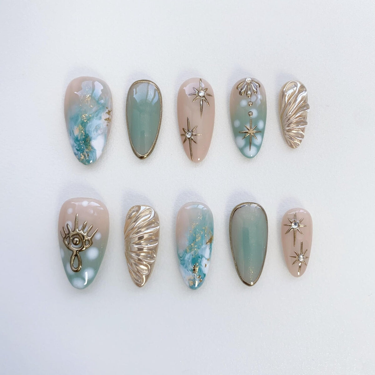 Set of ten false nails with a variety of designs. The nails are oval shaped and have a light pink and blue ombre effect. Some nails feature gold accents, including stars, seashells, and an eye design.