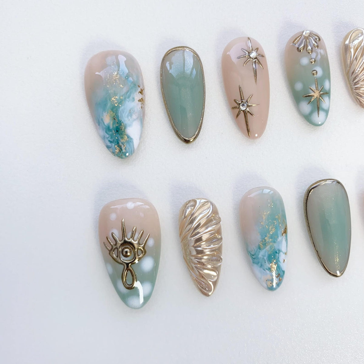 Set of ten false nails with a variety of designs. The nails are a light pink with a green ombre effect. Some nails feature gold accents, including stars, a seashell, and an eye.