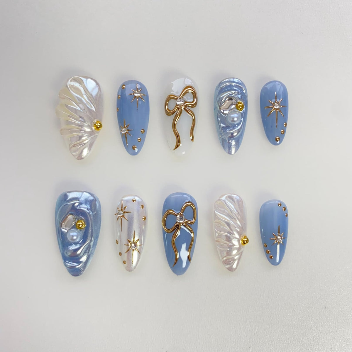Set of ten blue and white press-on nails with gold accents. The nails feature a variety of designs, including bows, stars, and pearls.