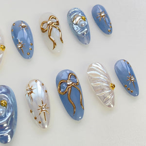 Set of ten blue and white oval-shaped fake nails with gold accents. Some nails feature a gold bow, others have a pearl and gold accents, and some have a gold star design.