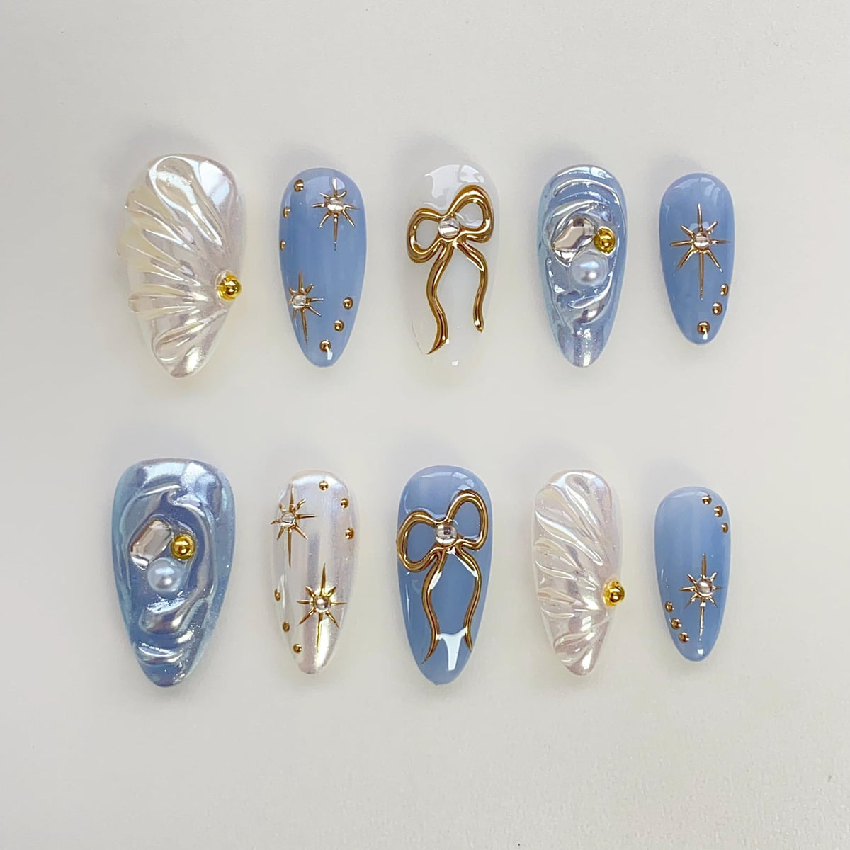 Set of ten blue and white press-on nails with gold accents. The nails feature a variety of designs including bows, stars, and pearl accents.