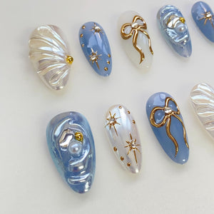 Set of ten false nails with various designs. Some nails are blue with gold accents, others are white with gold accents, and some are iridescent with pearl accents.