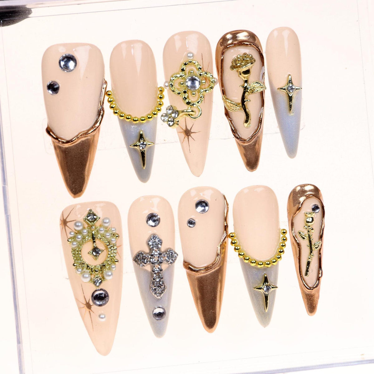 Set of ten false nails with a nude base and gold accents. Each nail features a different design, including a cross, a flower, a chain, and rhinestones.