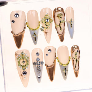 Set of ten false nails with a nude base and gold accents. Each nail features a different design, including a cross, a flower, a chain, and rhinestones.