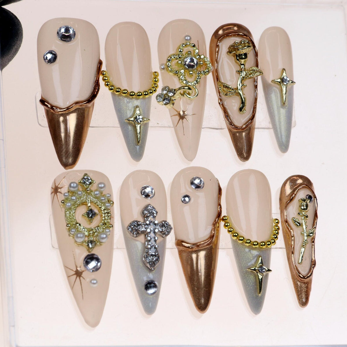 Set of ten false nails with a nude base and gold accents. Each nail features a different design, including a cross, a rose, and a chain.