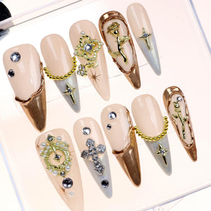 Set of ten false nails with a light pink base and gold accents. Each nail features a different design, including a cross, a rose, and a pearl-encrusted design.