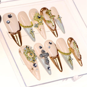Set of ten long, almond-shaped, light pink false nails with gold accents. Each nail is decorated with rhinestones, pearls, and gold charms, including crosses, stars, and a rose.