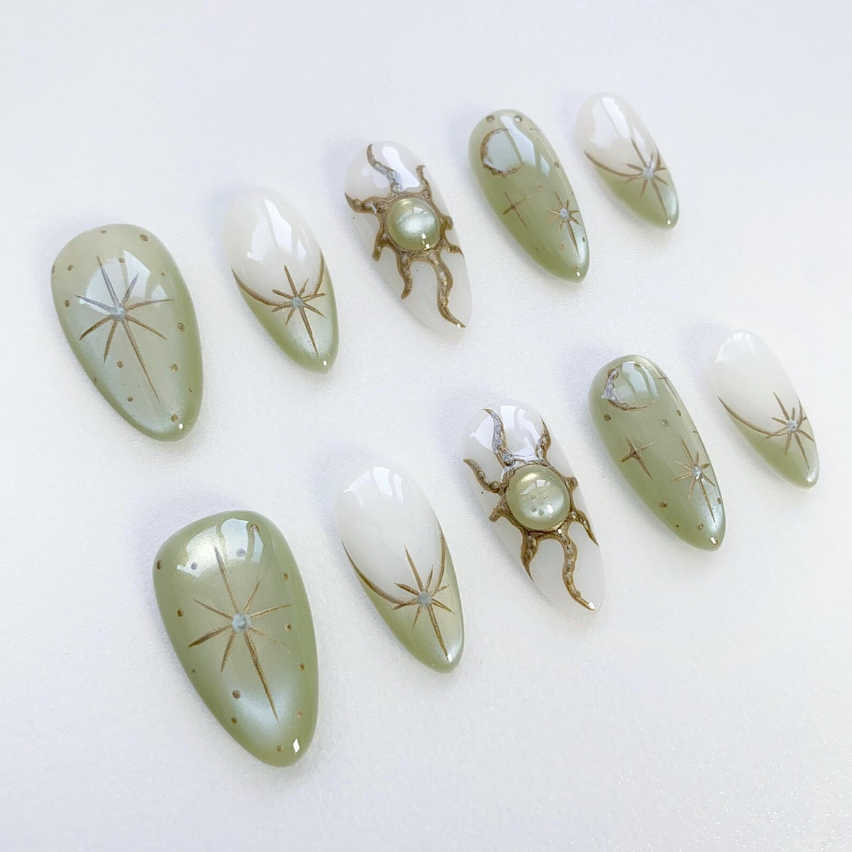 Set of ten green and white almond-shaped press-on nails with gold star and sun designs.