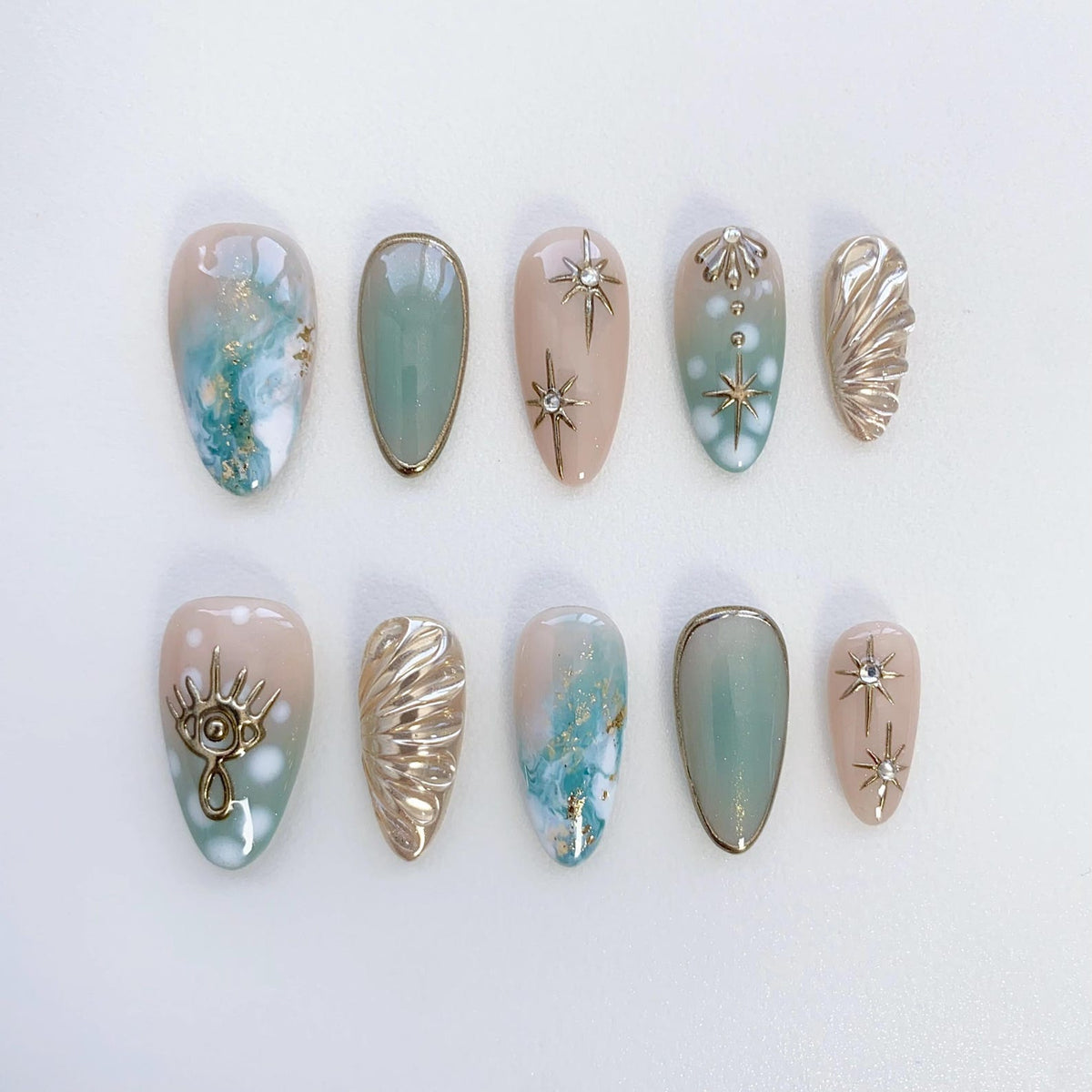 Ten press-on nails with a variety of designs. The nails are a light blue and beige color with gold accents. Some nails feature a gold star, a gold eye, and a gold shell design.