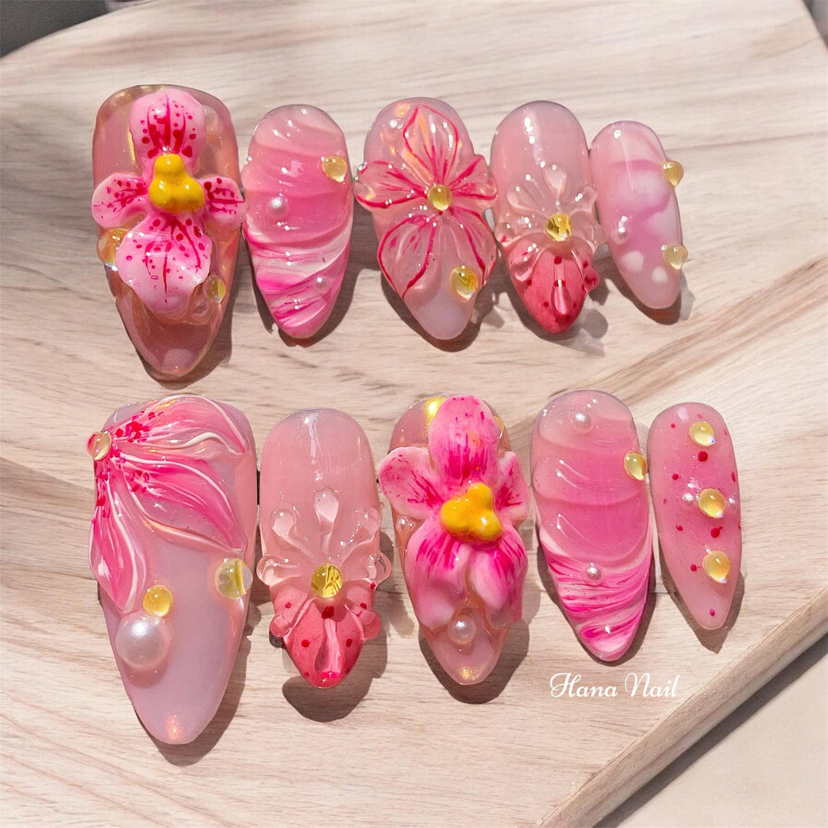 Pink and white oval-shaped fake nails with 3D floral designs. Some nails have small pearl accents. The text "Hana Nail" is visible in the bottom right corner of the image.