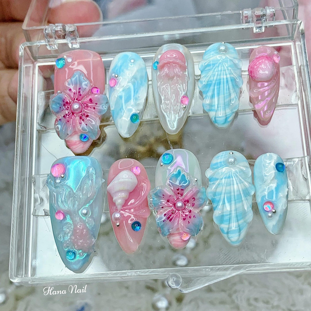 A set of ten false nails with a variety of ocean-themed designs. The nails are decorated with seashells, flowers, and swirls in shades of blue, pink, and white. The nails are displayed in a clear plas