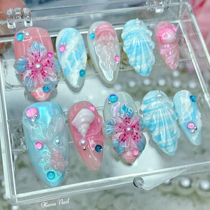 A set of ten false nails with a variety of ocean-themed designs. The nails are in a clear plastic box. The designs include a seashell, a jellyfish, a flower, and a mermaid.