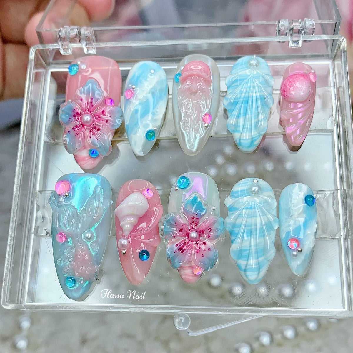 A set of ten false nails with various designs inspired by the ocean. The designs include seashells, flowers, and swirls. The nails are in a clear plastic case with the text "Hana Nail" printed on the 