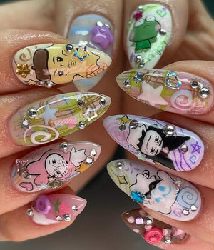 Close-up of ten fingernails with different designs. Each nail is painted with a clear base and decorated with various cartoon characters, glitter, and rhinestones. The designs include a frog, a cartoon character with a hat, a cartoon character with a