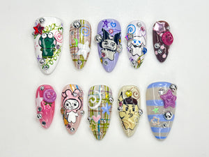 Set of ten false nails with various designs. The designs include a frog, a plaid pattern, a cartoon character, a swirl pattern, a striped pattern, and a flower. Each nail is decorated with rhinestones and other embellishments.