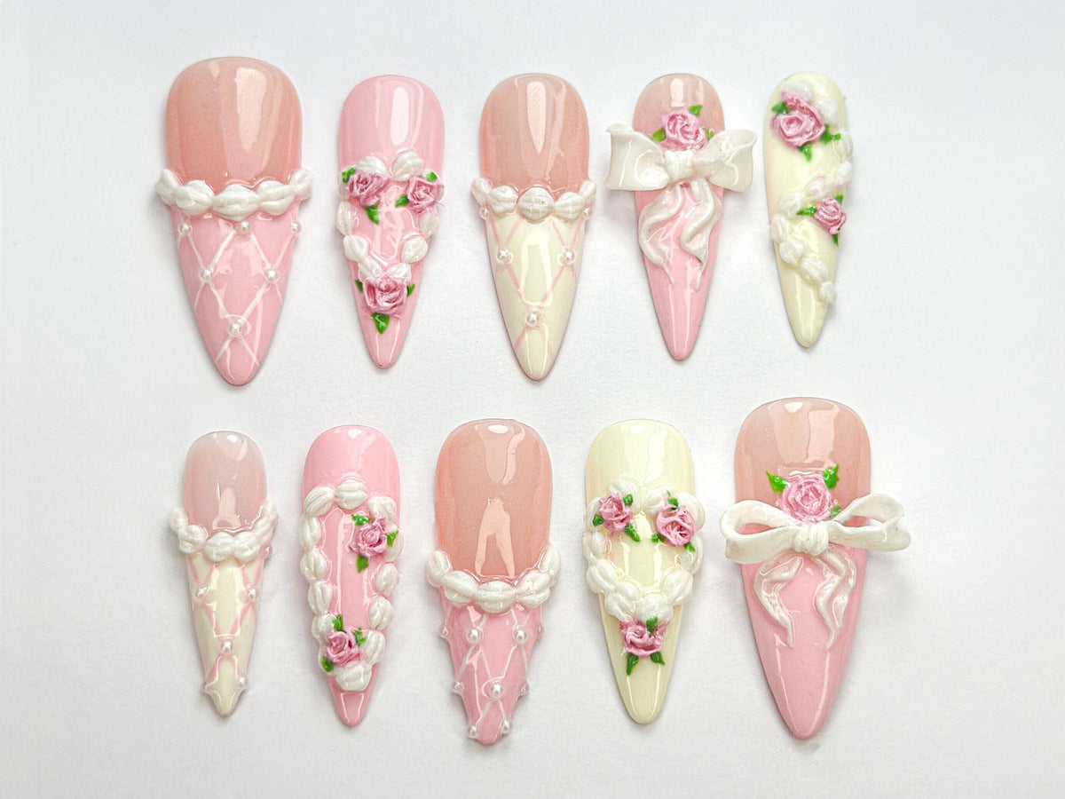 Ten pink and white false nails with different floral and ribbon designs. The nails are long and pointed.