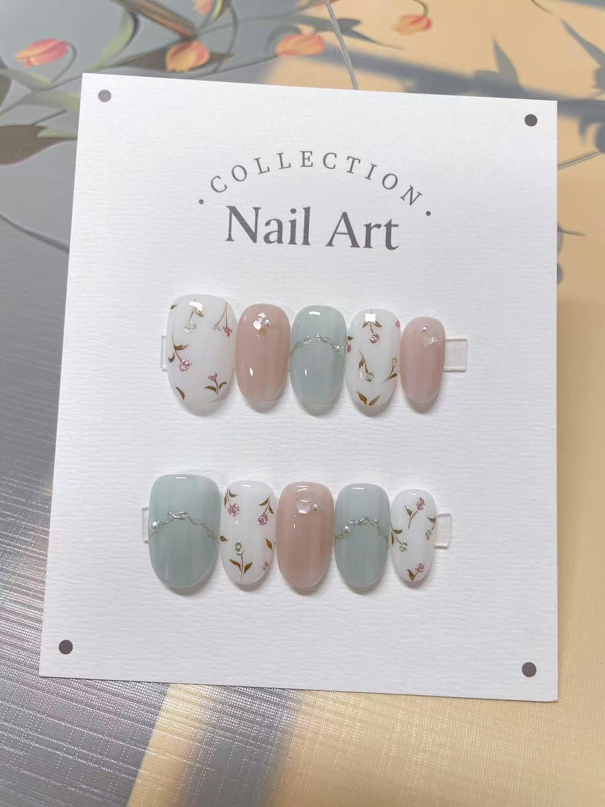 A set of nail art on a white card. The nails are oval-shaped and feature a variety of designs, including solid colors like pink and light blue, and floral patterns with small pink flowers and green st