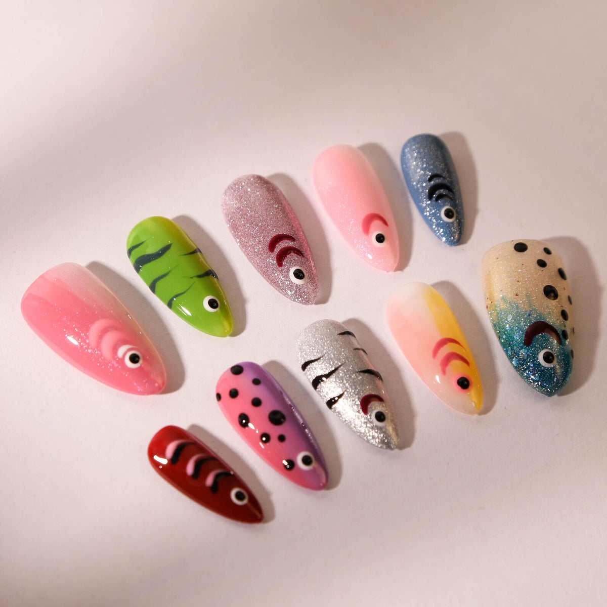 A collection of ten artificial nails, each uniquely designed with a fish theme. The nails feature various colors, patterns, and textures, including glitter, stripes, and polka dots. Each nail has a sm