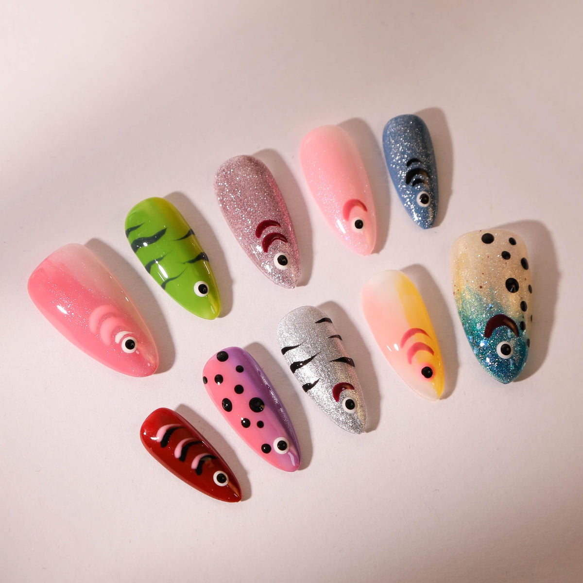 A collection of ten artificial nails, each uniquely designed with a fish theme. The nails feature various colors, including pink, green, silver, and blue, with detailed fish-like patterns and accents.
