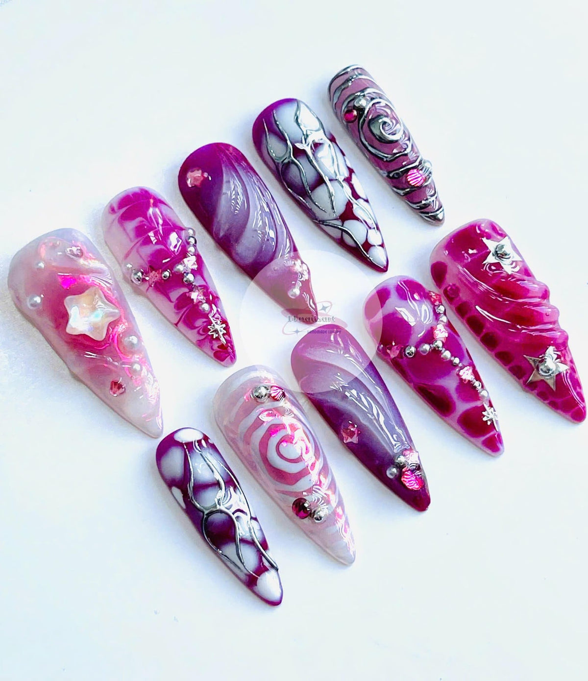 A collection of ten long, almond-shaped artificial nails. Each nail features a unique design with shades of pink, purple, and white, adorned with glitter, gems, and intricate patterns. Some nails have