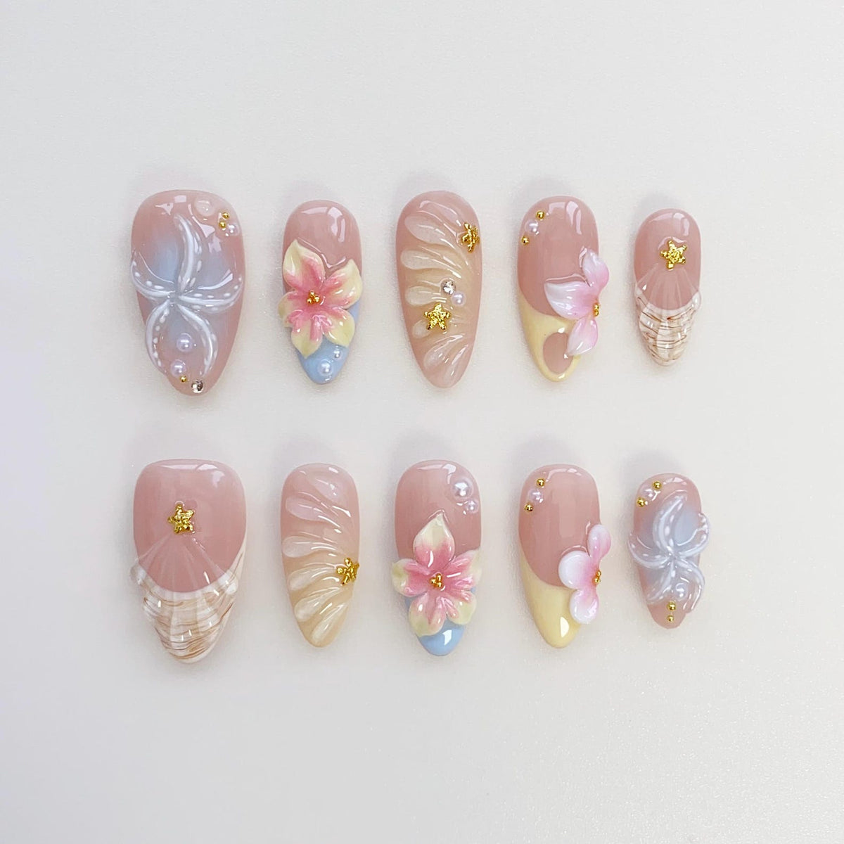 A set of ten artificial nails in various designs. Each nail is a light pink color with detailed floral and abstract designs. Some nails feature 3D flowers in pink, yellow, and blue, while others have 
