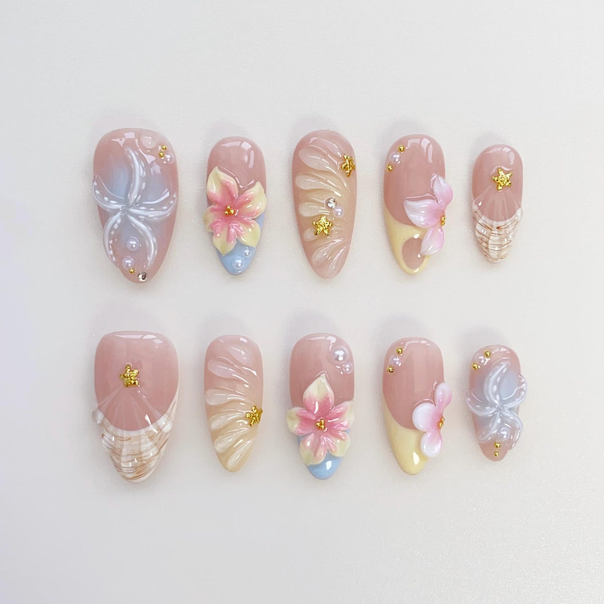 A set of ten artificial nails in various designs. The nails feature a light pink base with floral and abstract designs in white, blue, and pink. Some nails have 3D flower accents and gold embellishmen