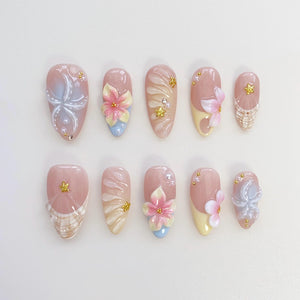 A set of ten artificial nails in various designs. The nails feature a light pink base with floral and abstract designs in white, blue, and pink. Some nails have 3D flower accents and gold embellishmen