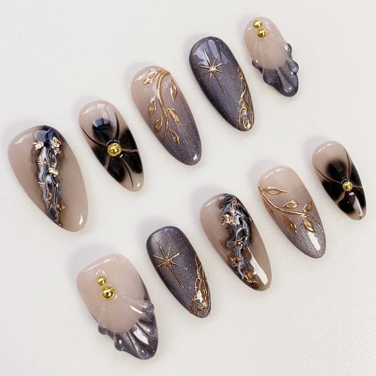 A set of ten artificial nails in various designs. The nails feature a neutral base color with accents of gold, black, and gray. Some nails have floral or starburst designs, while others have gold embellishments.