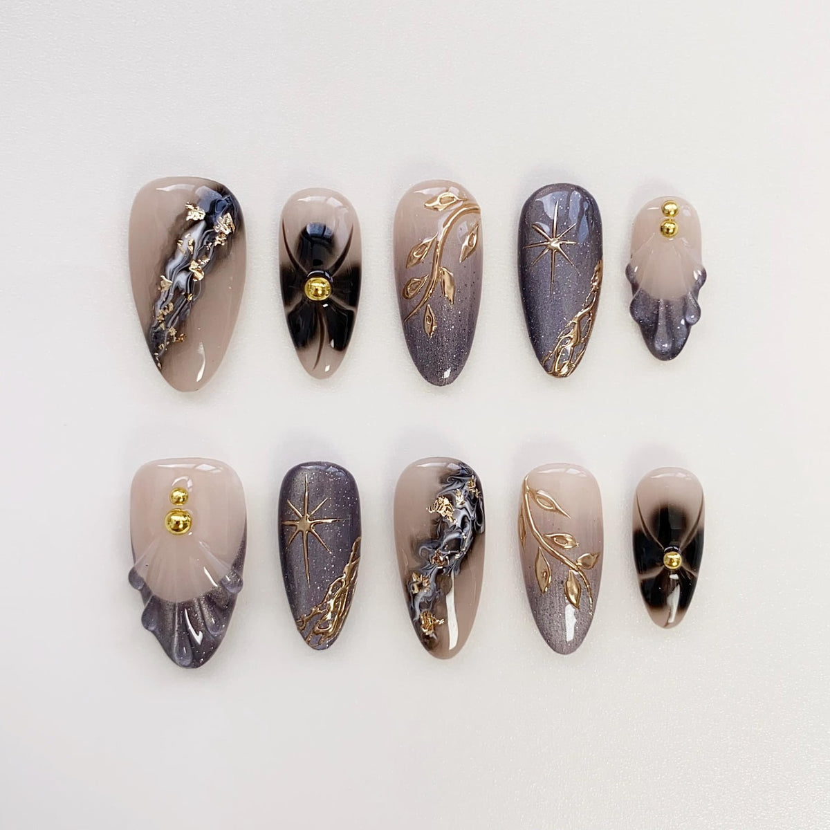 A set of ten almond-shaped false nails. The nails feature a variety of designs, including gold leaf accents, starbursts, and gradient effects in shades of beige, gray, and black. Some nails have gold embellishments.