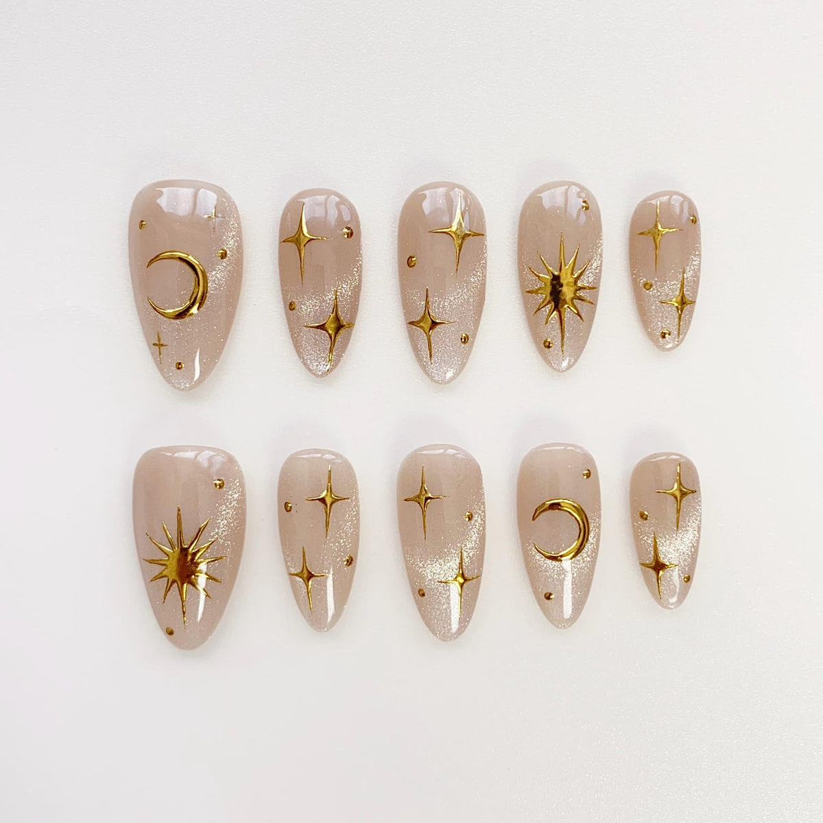 A set of ten almond-shaped false nails. Each nail is a nude color with gold celestial designs, including stars, moons, and suns. The nails have a glittery finish, adding a touch of sparkle.