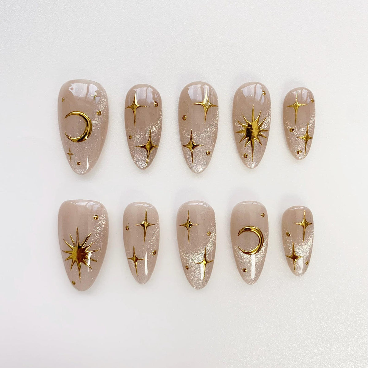 Set of ten almond-shaped false nails in a neutral beige hue, adorned with gold celestial designs. The designs include crescent moons, stars, and sunbursts, creating a cosmic theme. The nails have a glossy finish.