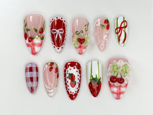 A set of ten almond-shaped false nails with various designs. The designs include strawberries, bows, and gingham patterns in red, white, and pink. Some nails feature 3D embellishments.