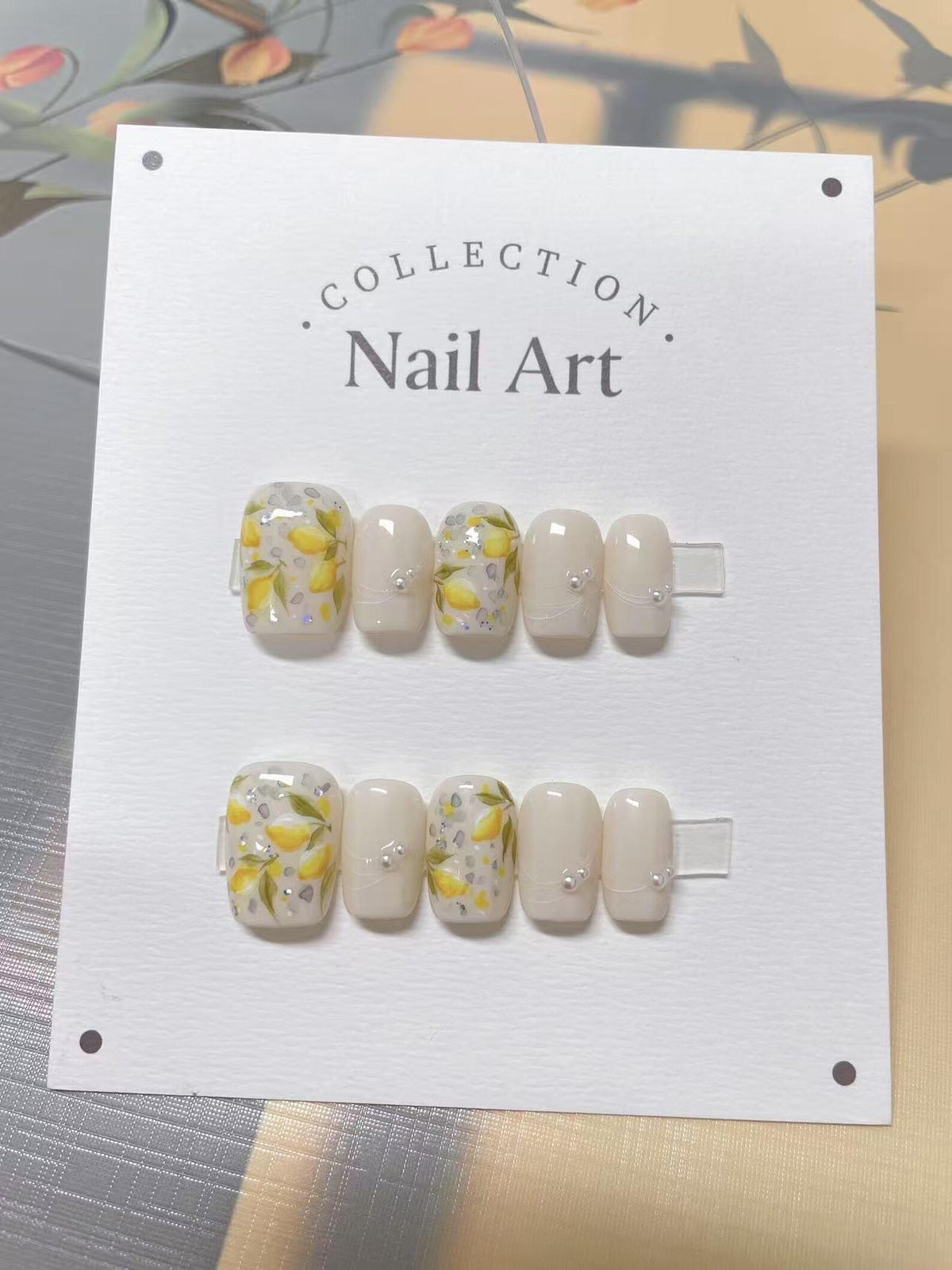 A set of 10 white press-on nails with a lemon and flower design. The nails are arranged in two rows of five on a white card with the text "COLLECTION. Nail Art."