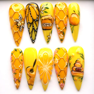 A set of ten long, yellow, almond-shaped artificial nails. Each nail features a unique design, including a bee, butterfly, honeycomb, honey pot with the word "HONEY", a pear, a guitar, and a flower. T