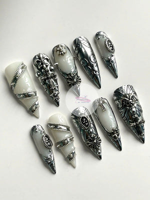 A collection of ten long, pointed false nails. Each nail features a silver metallic design with ornate details, including crosses, floral patterns, and geometric shapes. The base of each nail is a cre