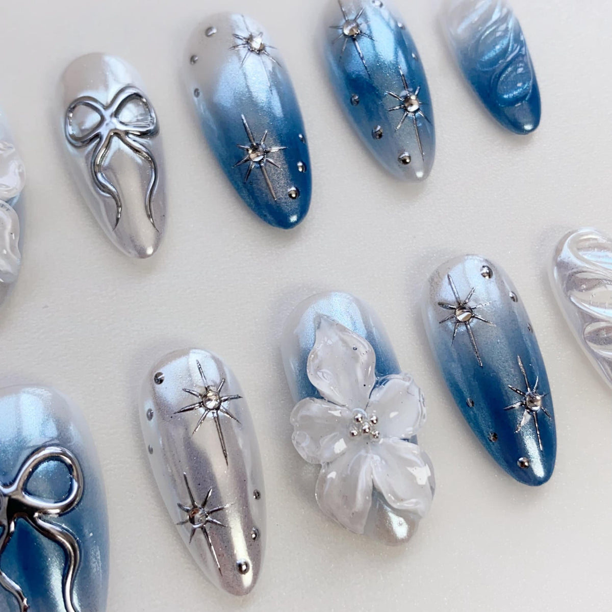 Oval-shaped artificial nails in various designs. Some nails feature a silver bow, others have a gradient of white to blue with star-shaped embellishments. One nail has a white flower with a silver center.