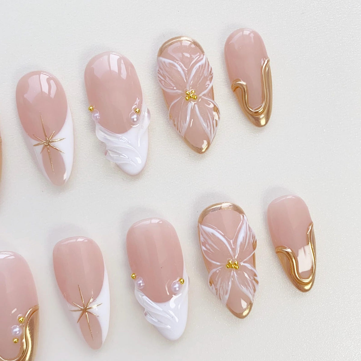 A collection of artificial nails in various designs. Some nails feature a nude base with white tips and gold star accents. Others showcase white floral designs, gold trim, and pearl embellishments. The nails are oval-shaped.