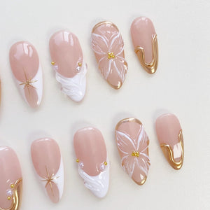 A collection of artificial nails in various designs. Some nails feature a nude base with white tips and gold star accents. Others showcase white floral designs, gold trim, and pearl embellishments. The nails are oval-shaped.