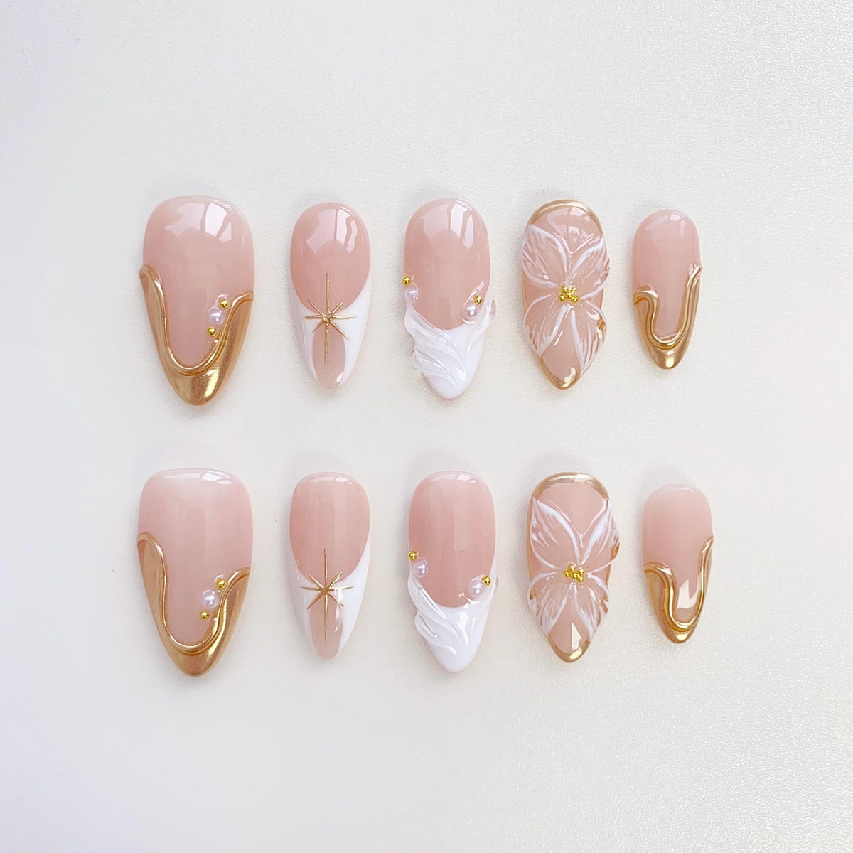 A set of ten almond-shaped false nails. The nails feature a pale pink base with gold accents, white designs, and floral embellishments. Some nails have a gold border, while others have a starburst or a flower design.