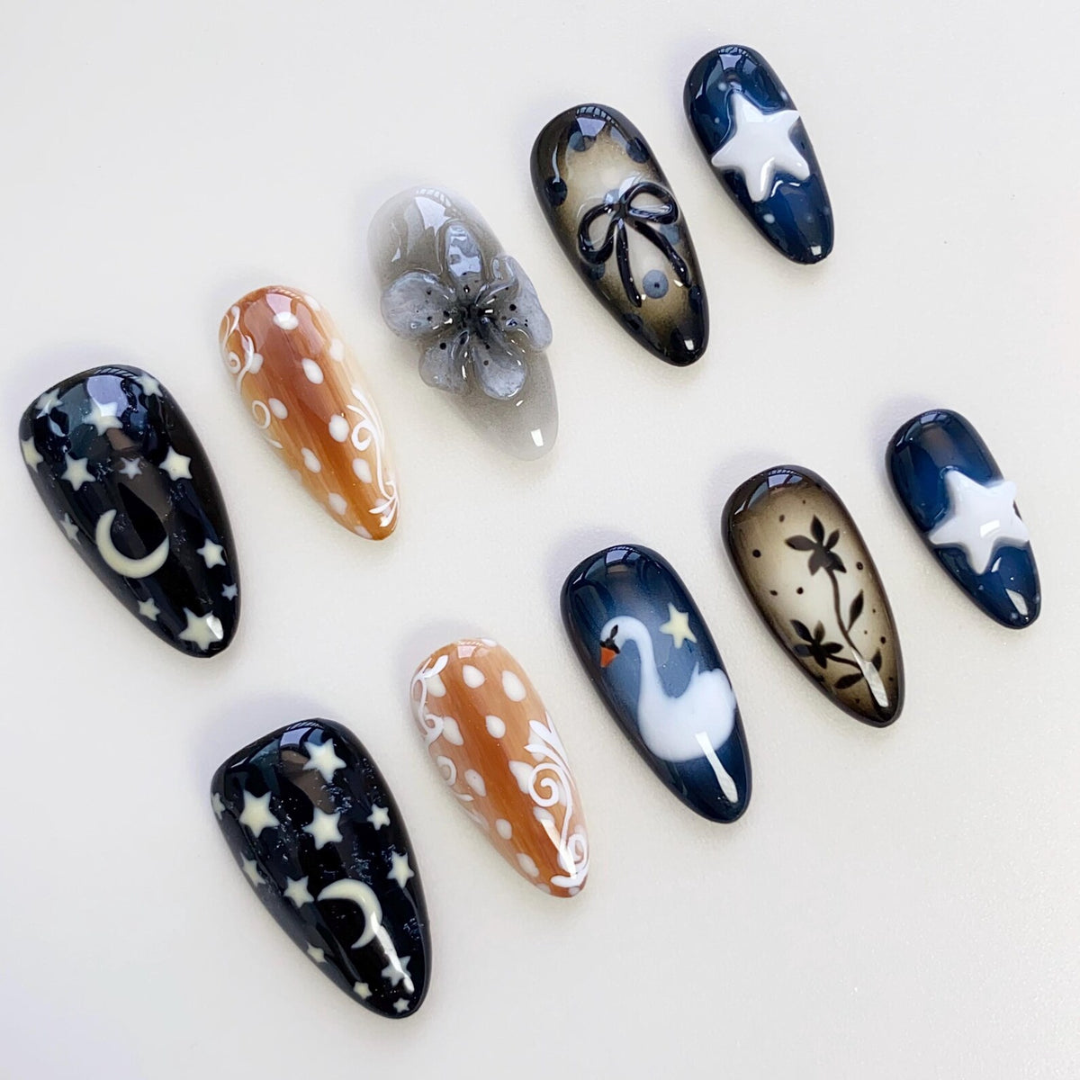 A collection of ten artificial nails with various designs. The nails feature a range of colors, including black, brown, and white. Designs include stars, moons, flowers, swans, and abstract patterns. The nails are oval-shaped and have a glossy finish