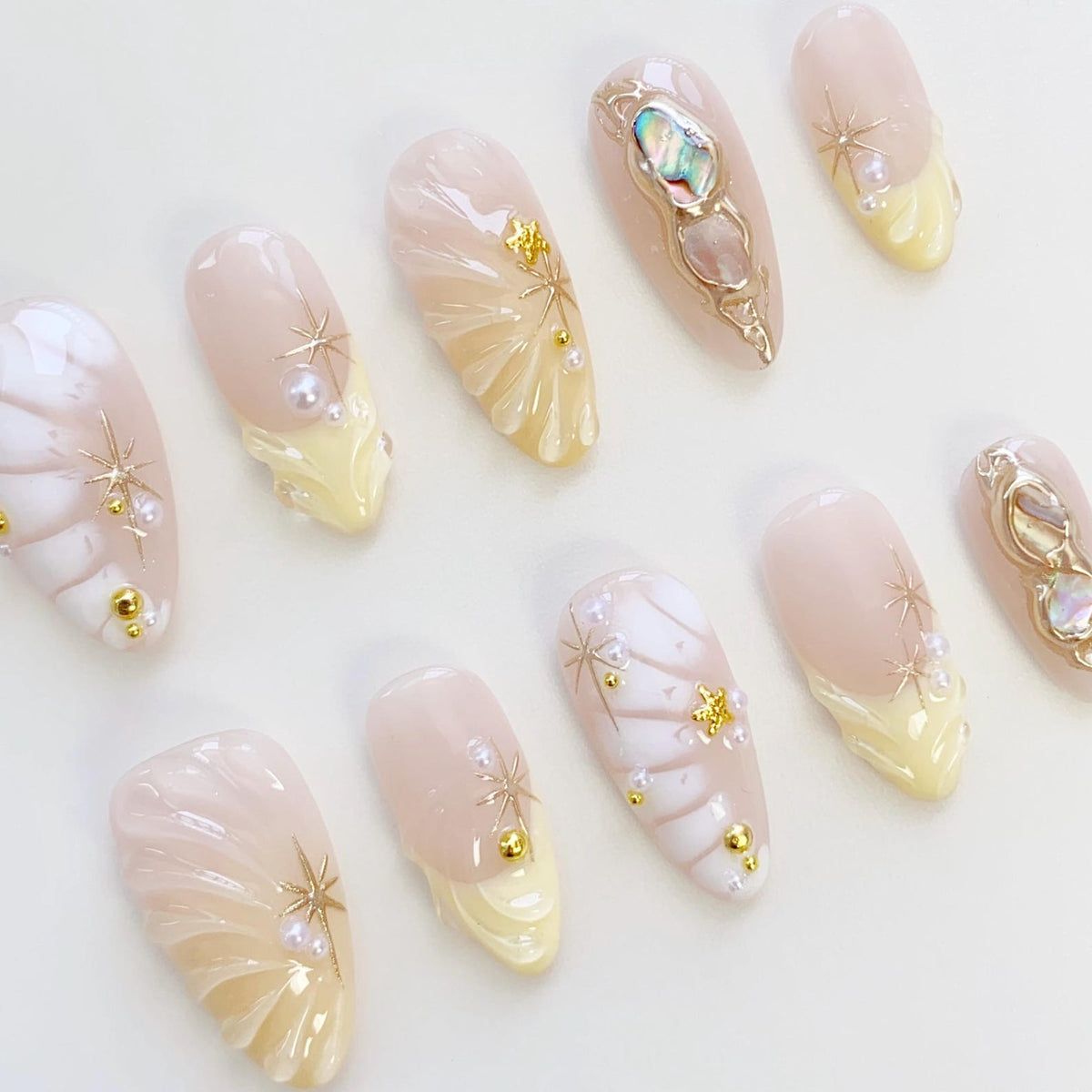 A set of ten almond-shaped artificial nails. Each nail features a light pink base with various designs, including gold star accents, pearls, and a yellow French tip. Some nails have iridescent embellishments.